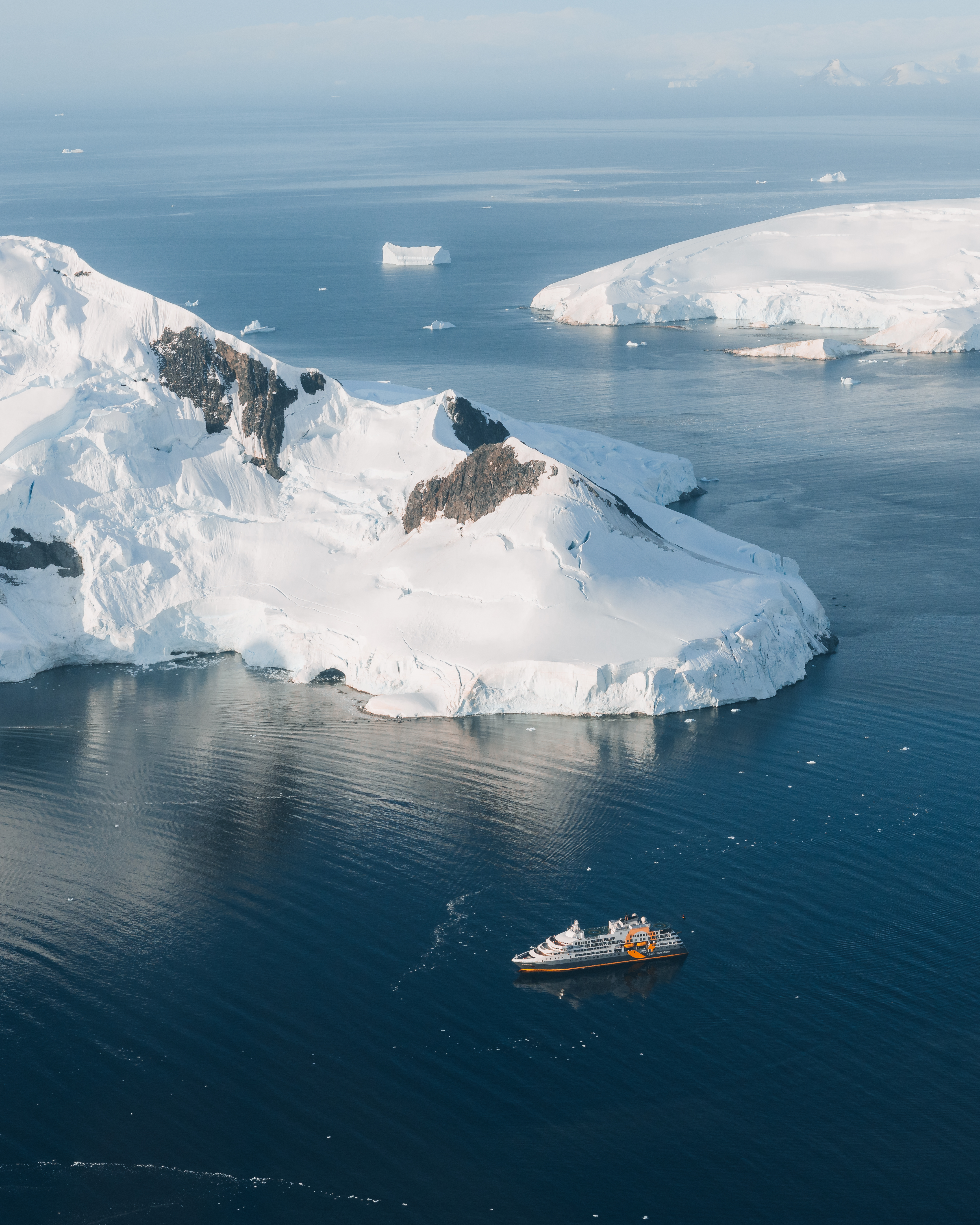 Expedition ship sailing below snow-covered Antarctic cliffs